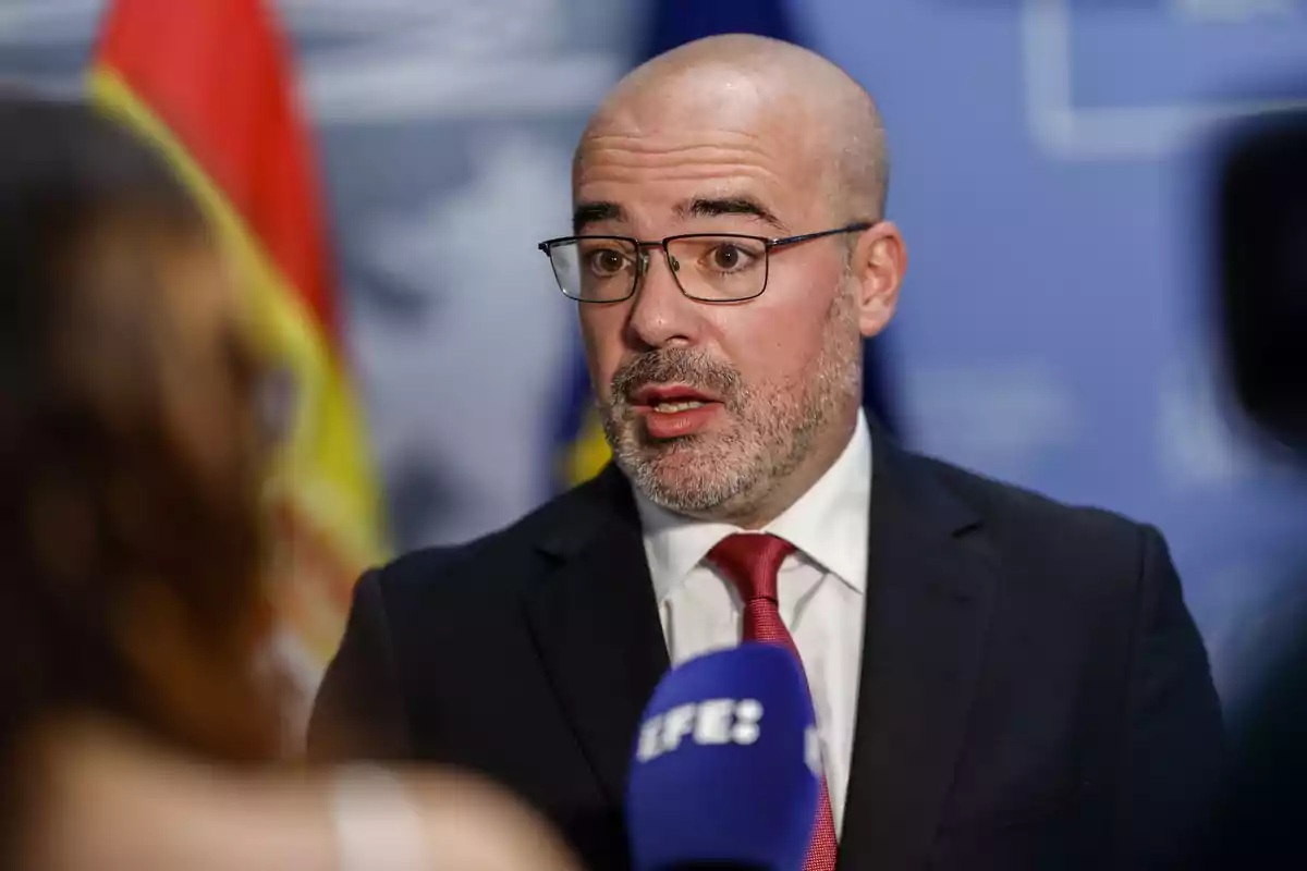 Un hombre con gafas y traje oscuro habla frente a un micrófono en un entorno formal.