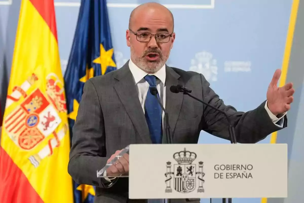 Un hombre en traje hablando en un podio con el escudo del Gobierno de España y banderas de fondo.