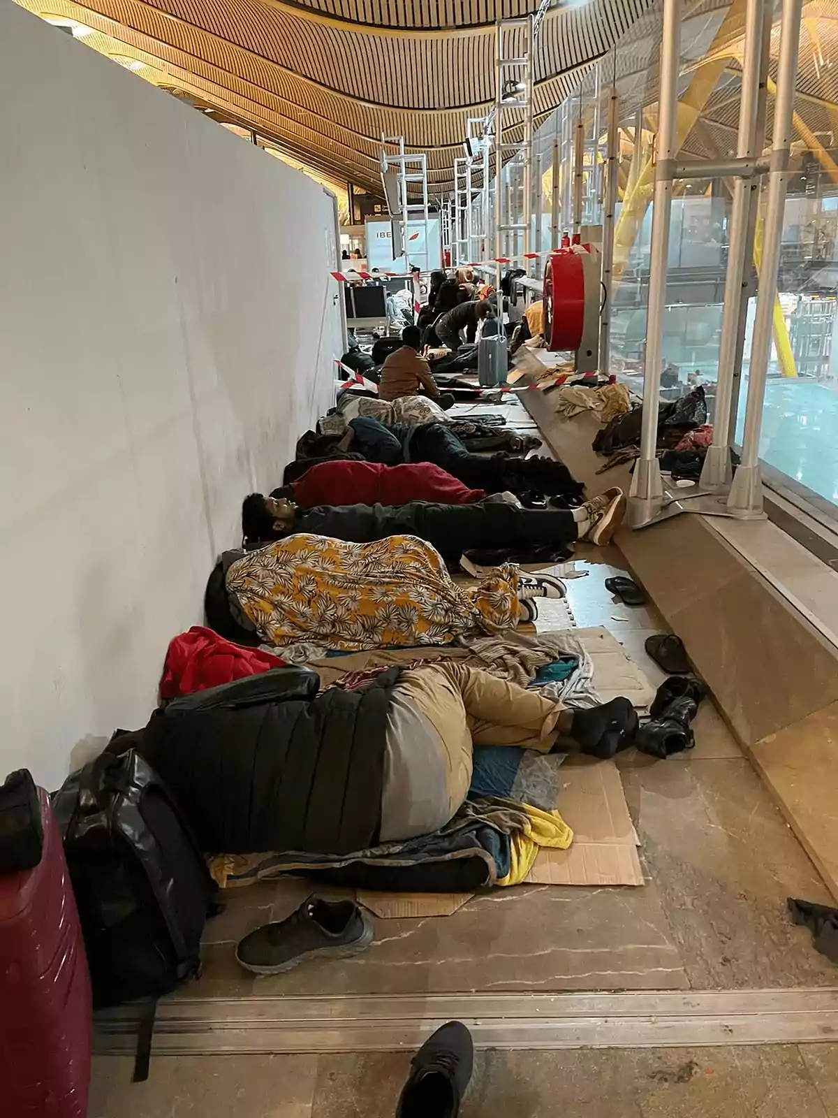 Personas durmiendo en el suelo de un aeropuerto, rodeadas de equipaje y cubiertas con mantas. Personas durmiendo en el suelo de un aeropuerto, rodeadas de equipaje y cubiertas con mantas.