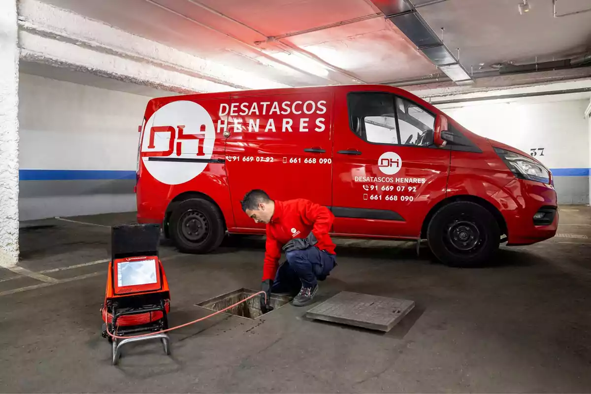 Un trabajador de Desatascos Henares inspecciona una alcantarilla en un estacionamiento subterráneo, junto a una furgoneta roja de la empresa equipada con herramientas especializadas.