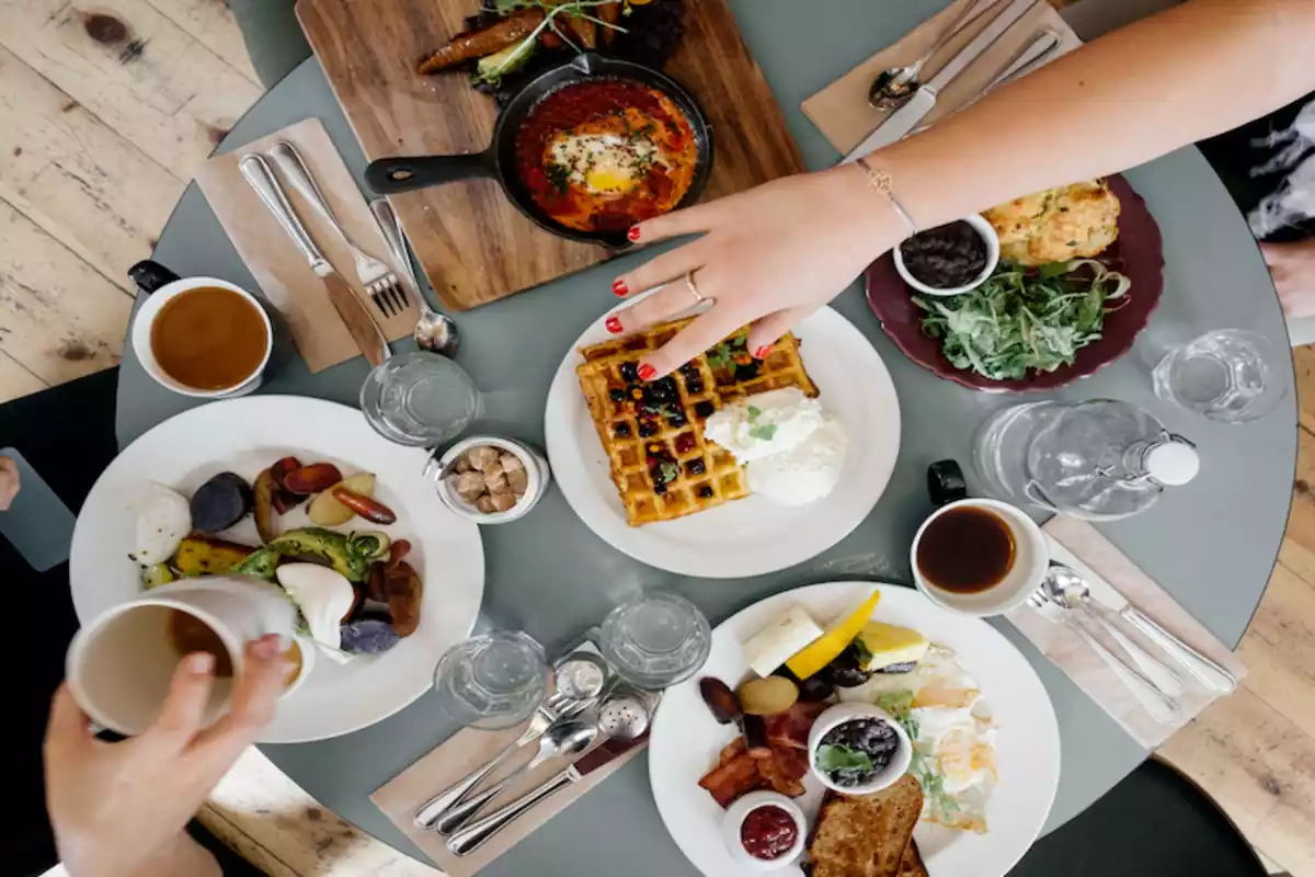 Dos personas comparten un desayuno de lo más contundente en una mesa redonda, con un gran gofre en el centro, cafés y otros alimentos como huevos y vegetales