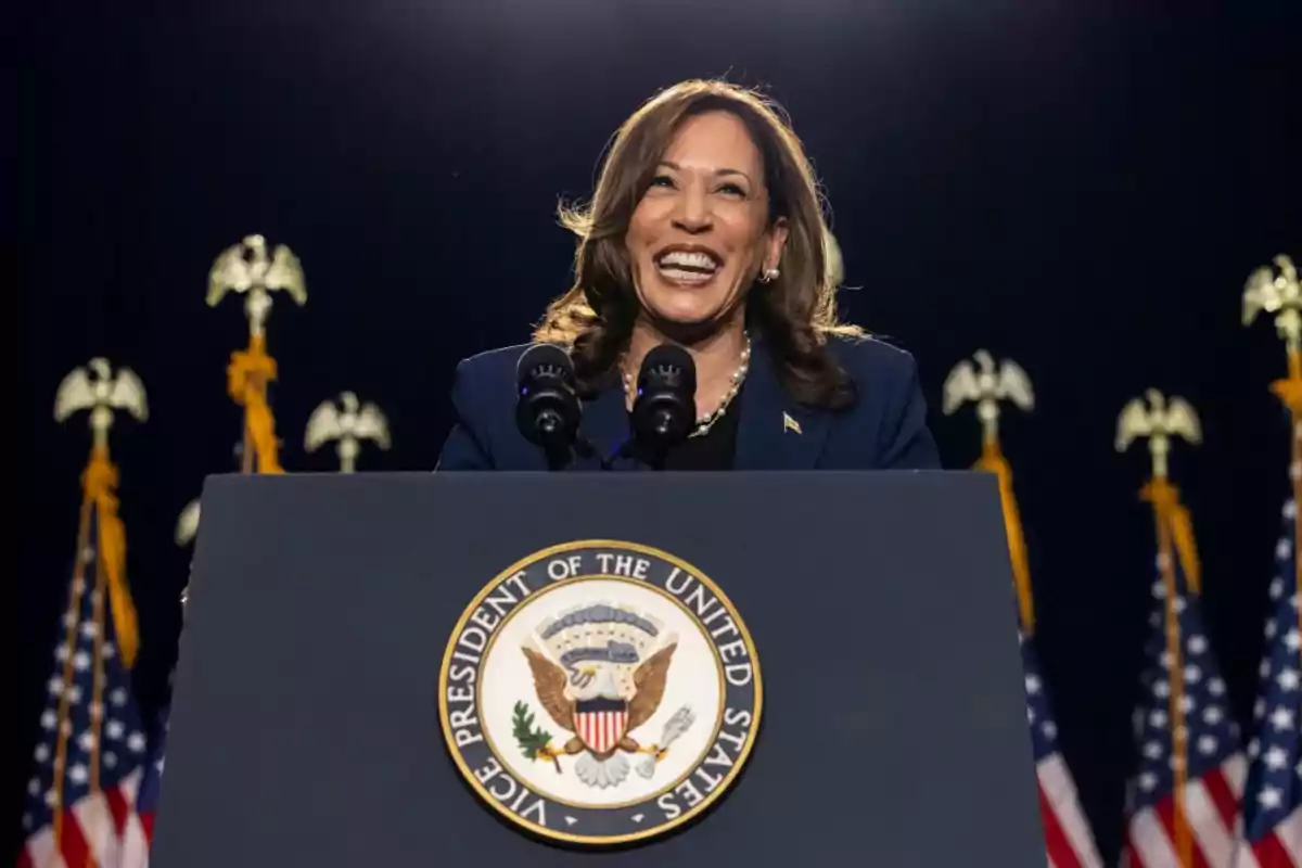 Una persona sonriendo detrás de un podio con el sello del Vicepresidente de los Estados Unidos y varias banderas estadounidenses en el fondo.