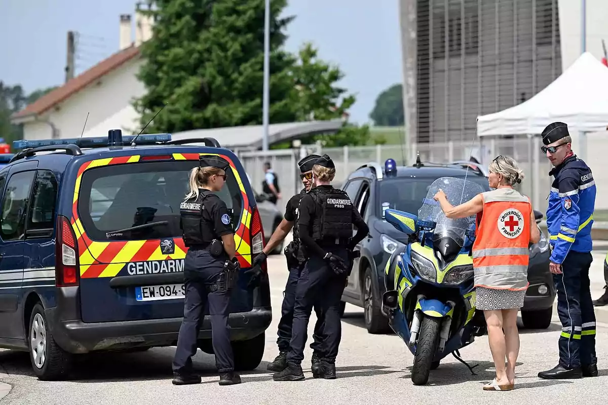 Varios agentes de la gendarmería francesa conversan junto a vehículos oficiales mientras una mujer con chaleco de la Cruz Roja Francesa señala algo en una motocicleta