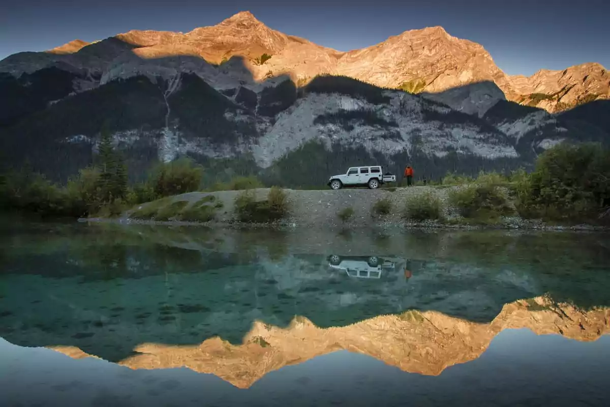 Un jeep blanco estacionado junto a un lago cristalino con montañas al fondo reflejadas en el agua y una persona de pie cerca del vehículo