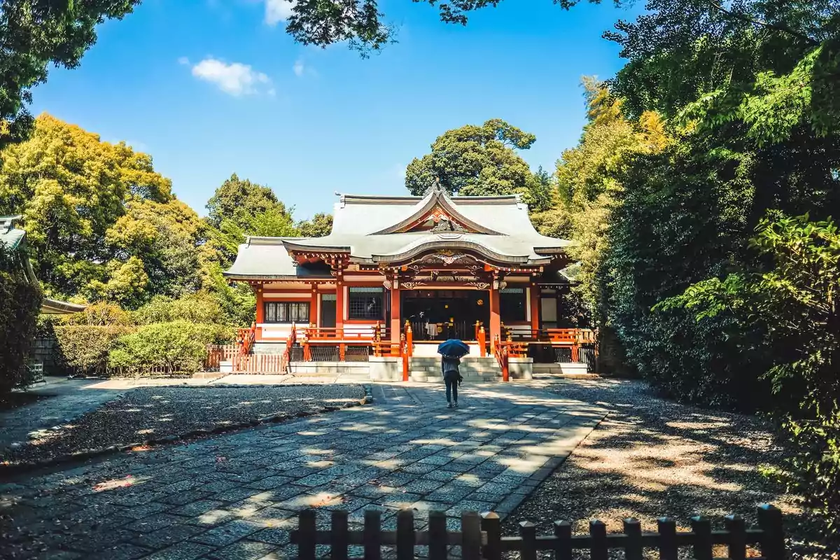 Un templo tradicional japonés rodeado de árboles con una persona caminando hacia él bajo un cielo azul.