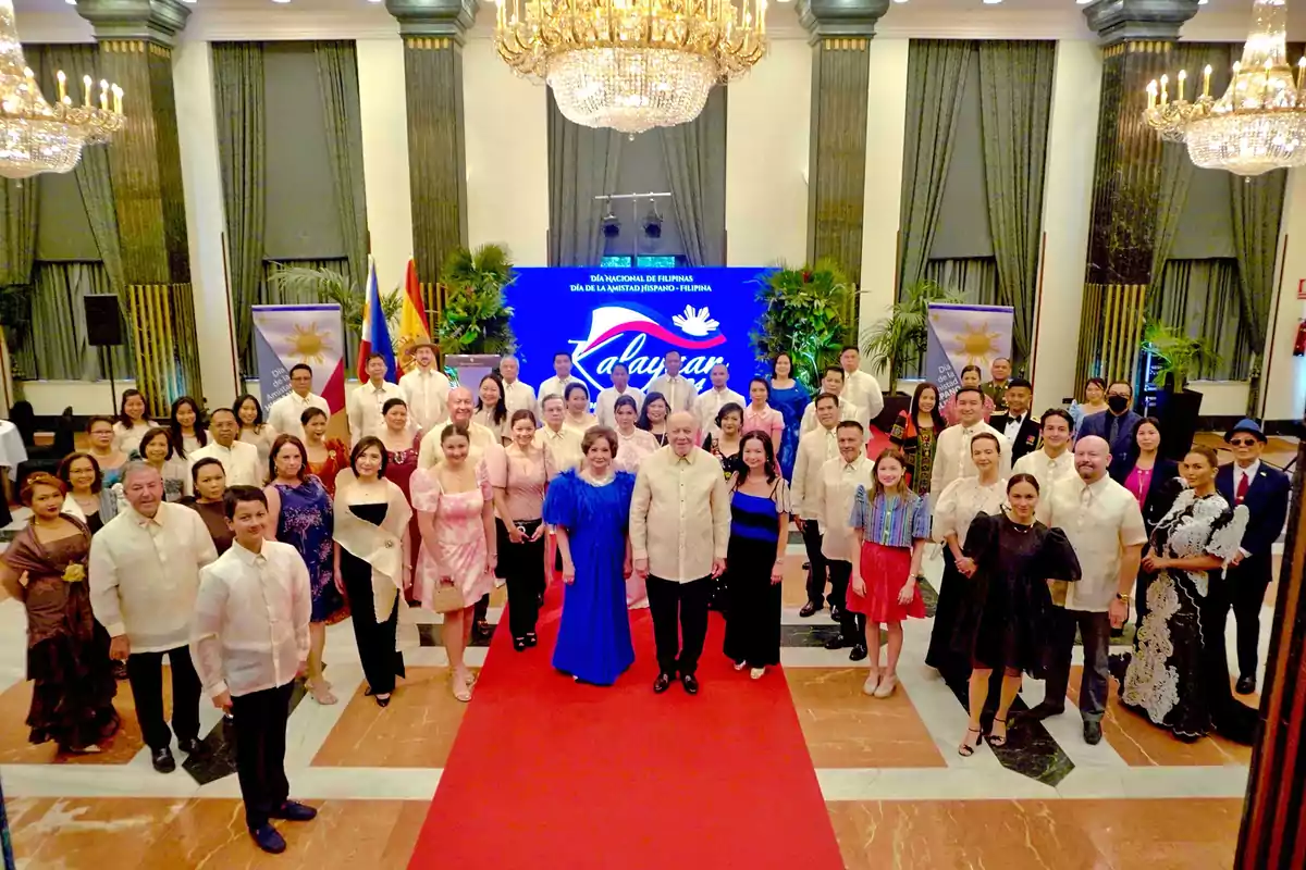 Un grupo de personas posando en un evento formal con una alfombra roja, candelabros y una pantalla azul de fondo que dice "Día Nacional de Filipinas, Día de la Amistad Hispano-Filipina".