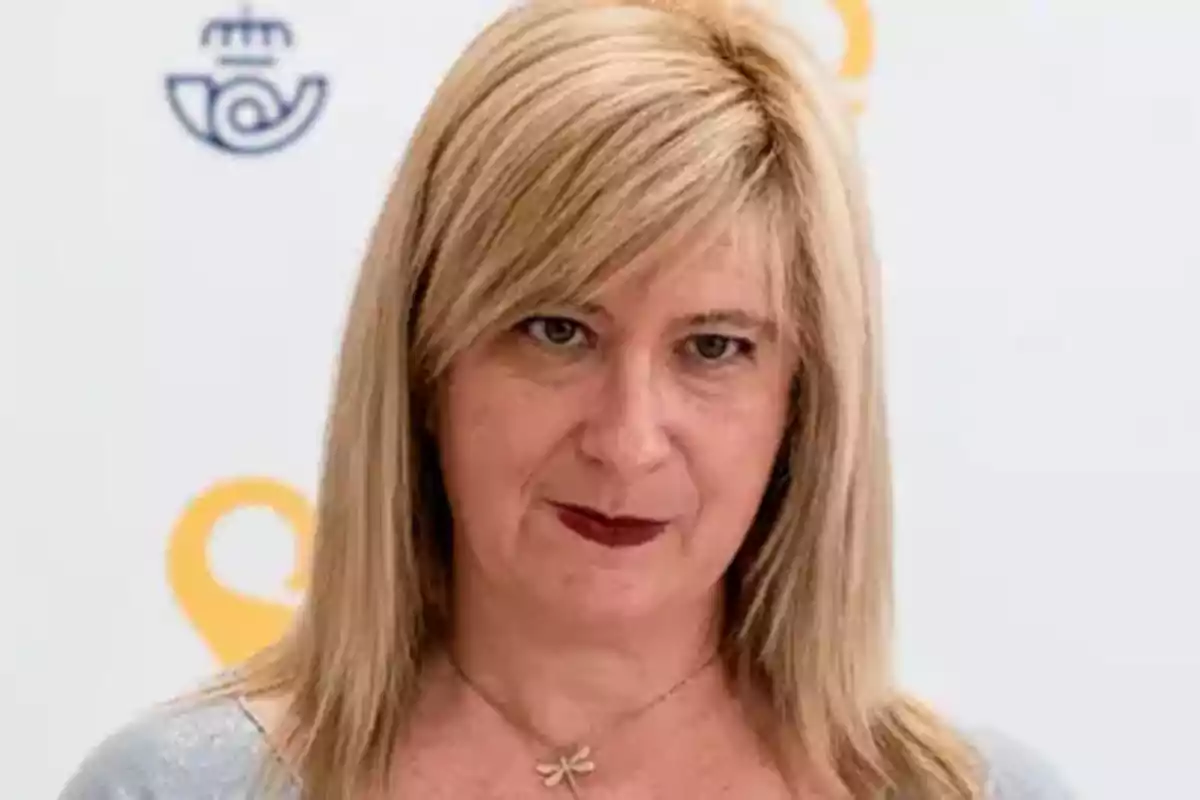 Una mujer rubia con cabello lacio y expresión seria lleva un collar con un colgante de libélula y está frente a un fondo blanco con logotipos. Una mujer rubia con cabello lacio y expresión seria lleva un collar con un colgante de libélula y está frente a un fondo blanco con logotipos.