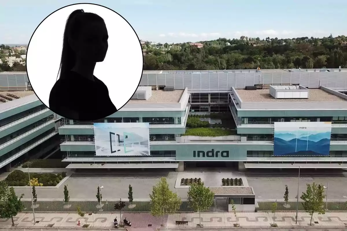 En la imagen se observa un edificio moderno con la palabra "Indra" en la fachada, lo que sugiere que es la sede de la empresa Indra. En la parte superior izquierda de la imagen, hay un círculo que contiene la silueta de una persona, pero no se puede identificar quién es debido a la falta de detalles. El entorno del edificio parece ser una zona urbana con vegetación y un camino peatonal en primer plano.