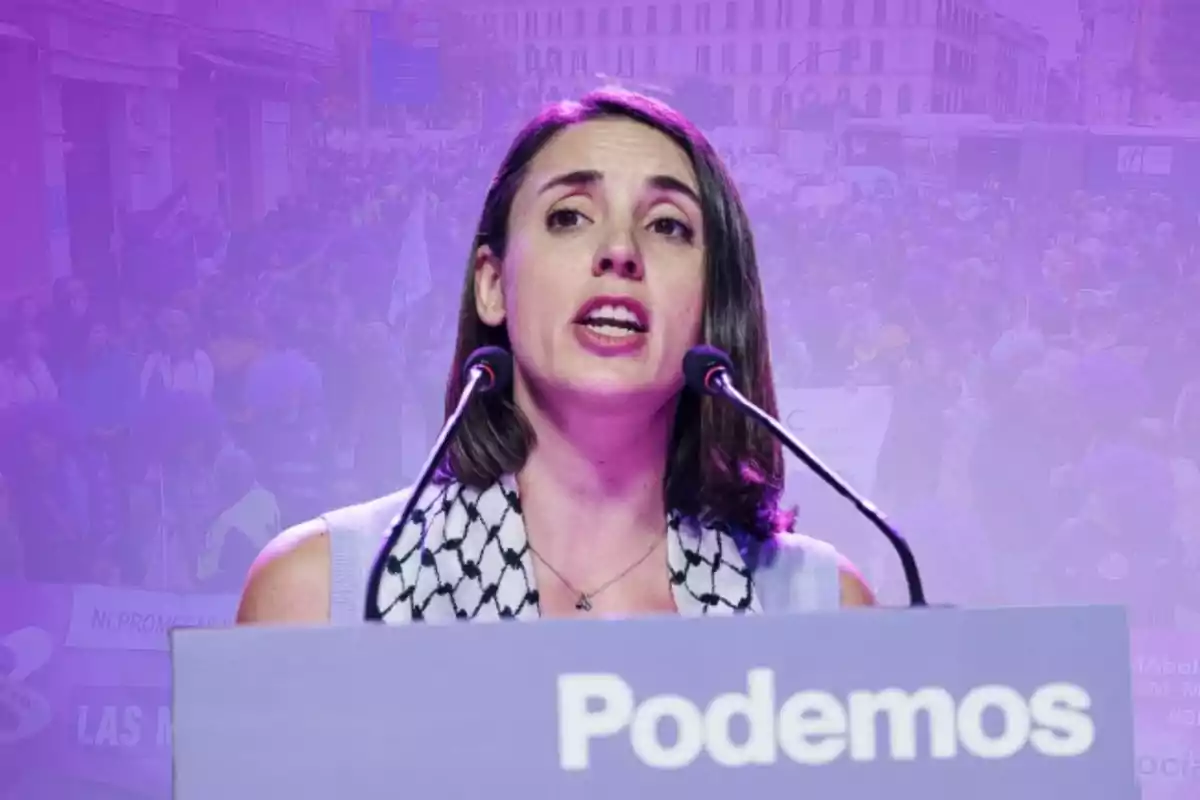 Una mujer hablando en un podio con un fondo morado y la palabra "Podemos" en el frente del podio.