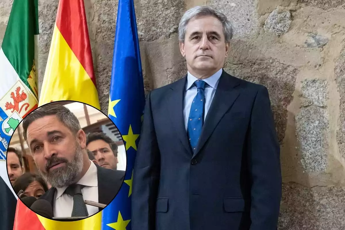 Un hombre de traje y corbata azul está de pie frente a una pared de piedra, con las banderas de Extremadura, España y la Unión Europea detrás de él; en la esquina inferior izquierda hay un círculo con la imagen de otro hombre con barba hablando frente a micrófonos.