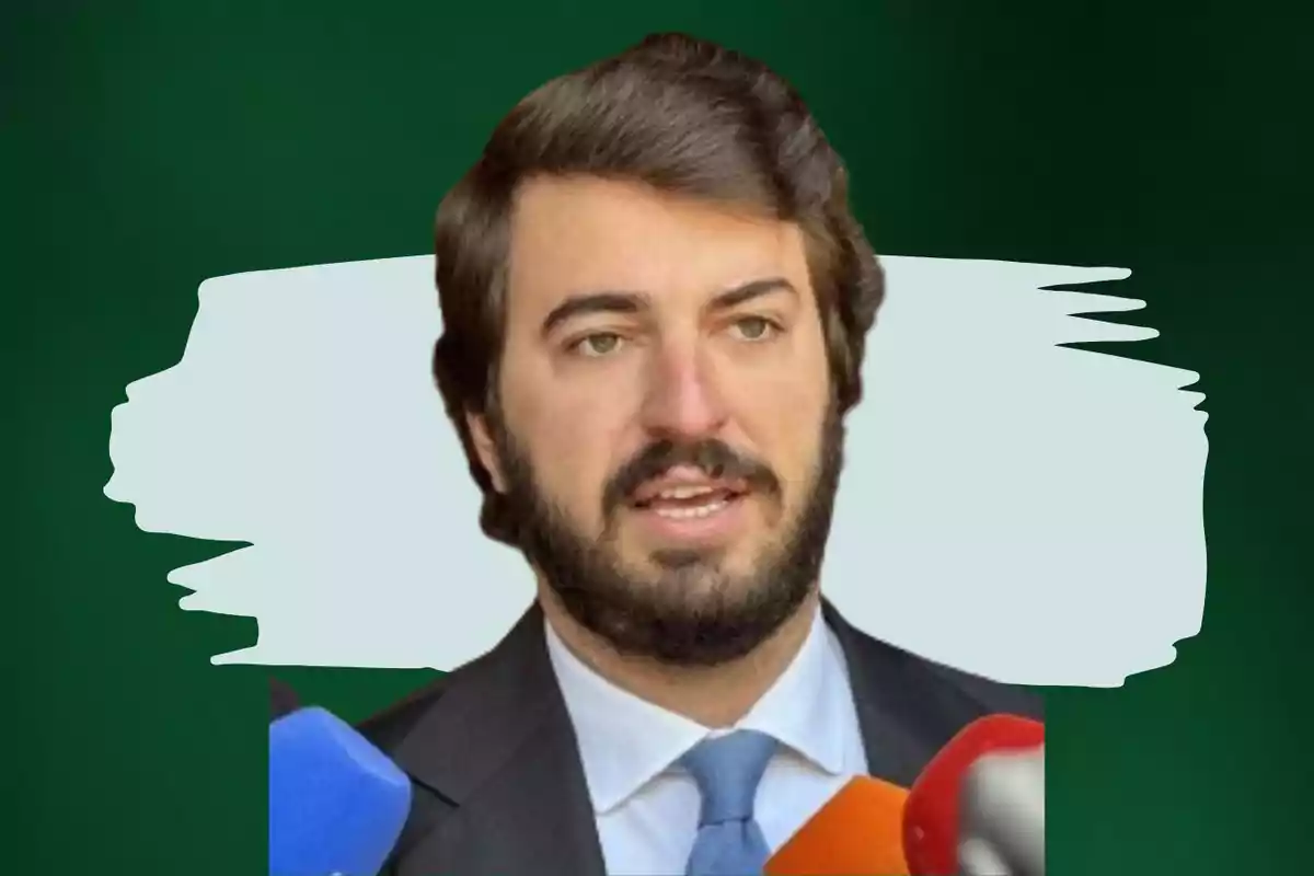 Hombre con barba y traje hablando frente a varios micrófonos con fondo verde y blanco.