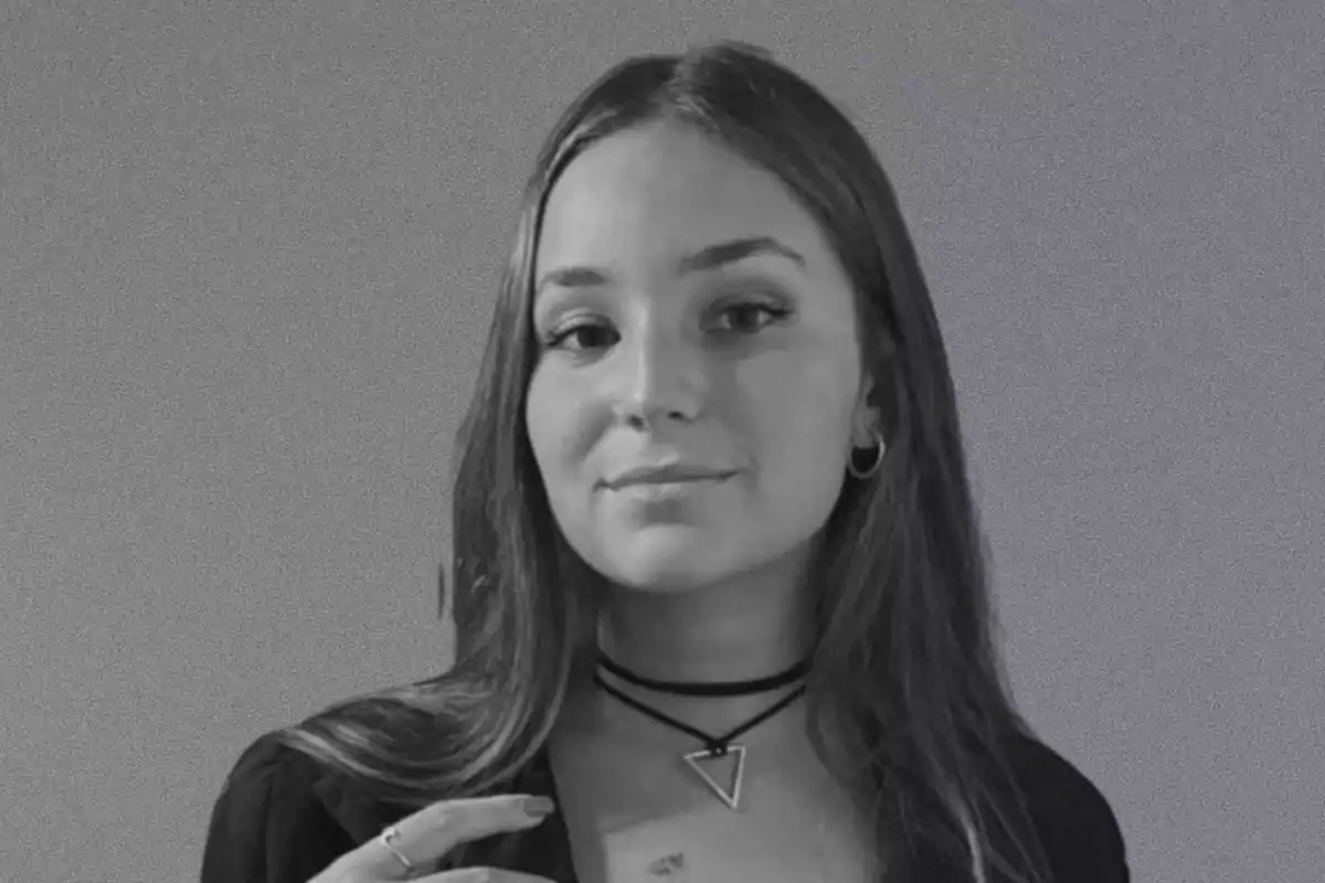 Una mujer con cabello largo y lacio, usando un collar con un colgante triangular, posando frente a un fondo gris.