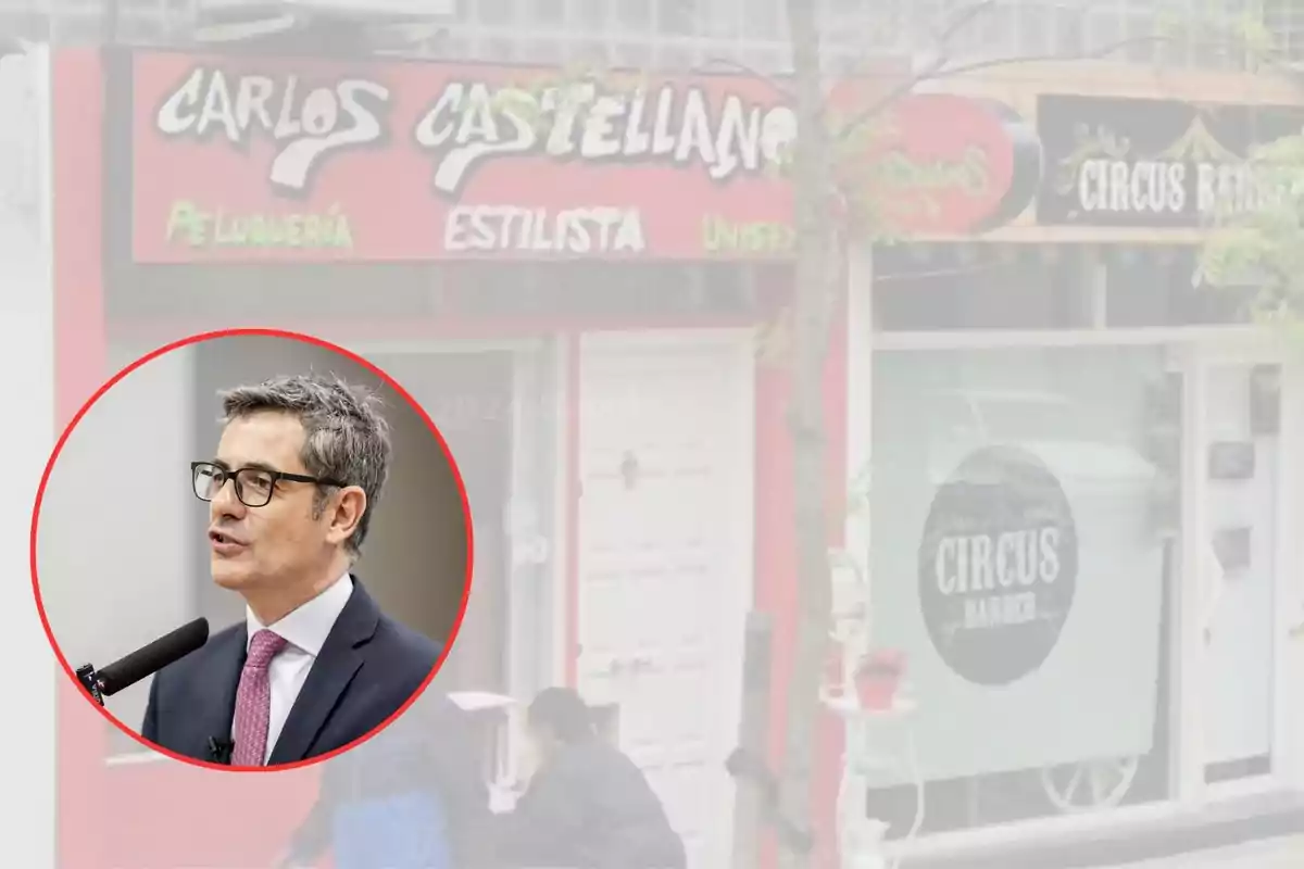 Un hombre con gafas y traje habla frente a un micrófono, con un fondo de una peluquería y barbería.