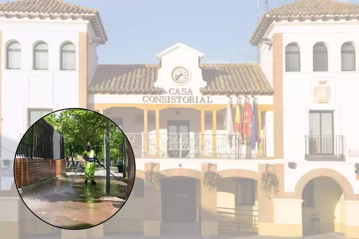 Un trabajador de limpieza con uniforme amarillo y verde está lavando una acera con una manguera de agua a presión, mientras que de fondo se observa la fachada de un edificio con el letrero "Casa Consistorial" y varias banderas.