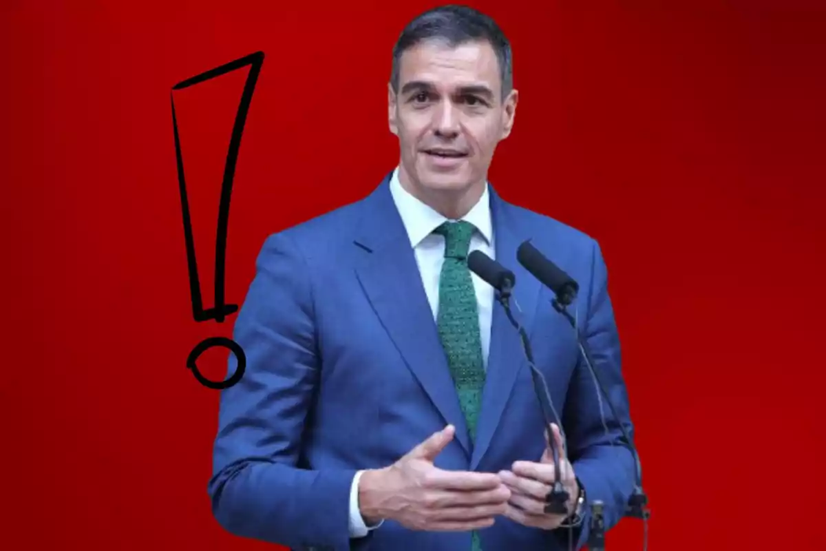 Hombre de traje azul y corbata verde hablando frente a dos micrófonos con un fondo rojo y un signo de exclamación negro a su izquierda.