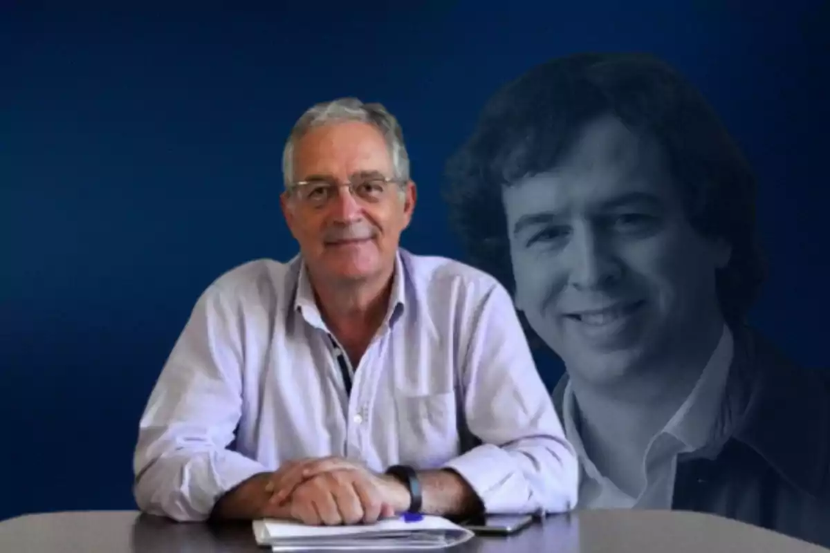 Un hombre mayor con camisa clara y gafas sentado en una mesa con un fondo azul y la imagen en blanco y negro de un hombre más joven detrás.