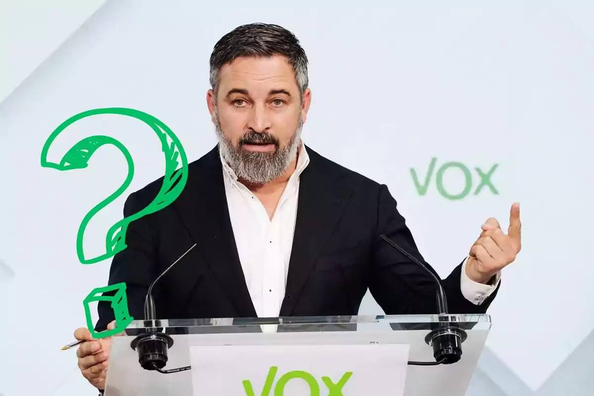 Un hombre con barba y traje oscuro hablando en un podio con el logo de VOX y un signo de interrogación verde a su izquierda.