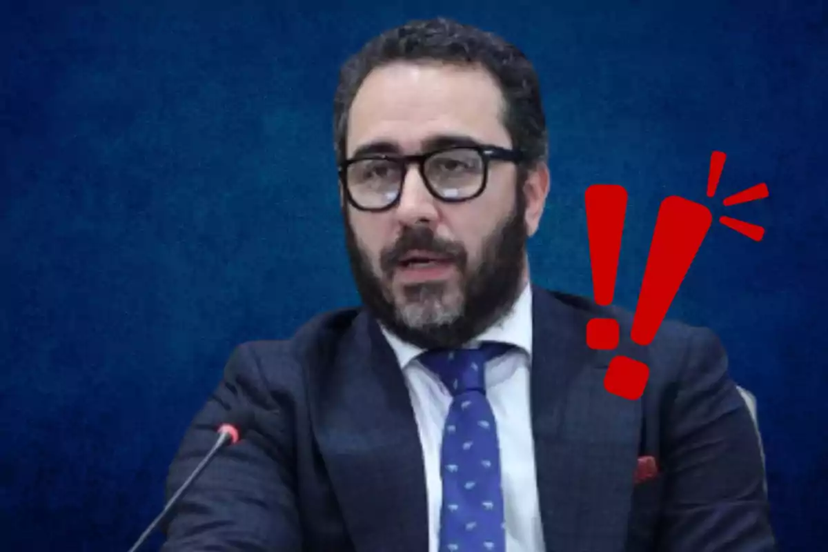 Hombre con barba y gafas en traje oscuro con corbata azul y un símbolo de exclamación rojo al lado sobre un fondo azul.