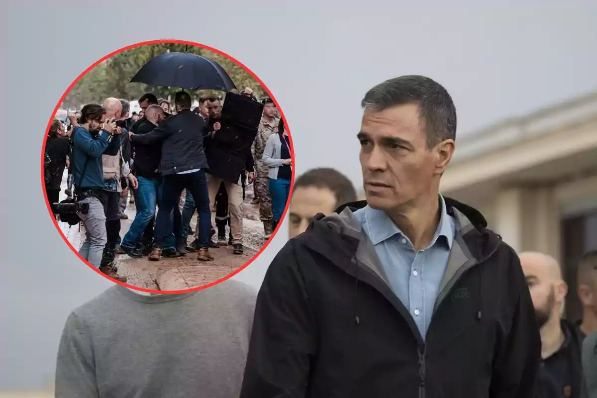 Un hombre con chaqueta oscura y camisa azul está de pie al aire libre mientras un grupo de personas con cámaras y paraguas se encuentra en el fondo.