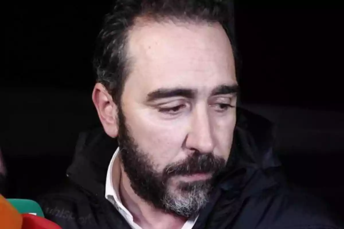 Hombre con barba y cabello oscuro mirando hacia abajo con expresión seria.