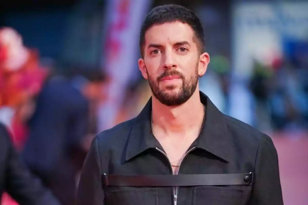 Un hombre con barba y cabello corto viste una chaqueta oscura en un evento con fondo desenfocado.