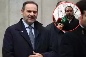 Un hombre con barba y traje oscuro está sonriendo mientras camina al aire libre, con un recuadro que muestra a otra persona siendo entrevistada por varios micrófonos.