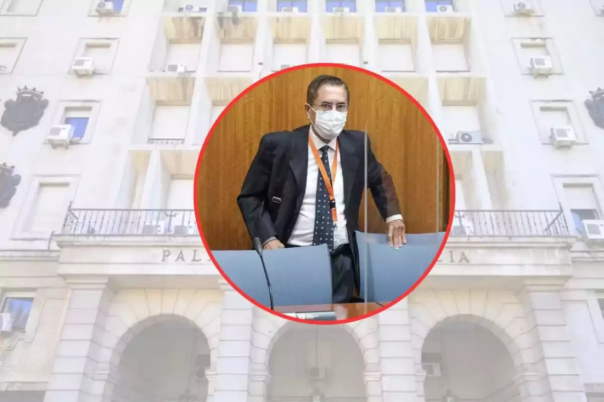 Un hombre con mascarilla y traje oscuro está de pie detrás de una silla en una sala de conferencias, con un edificio de fondo.