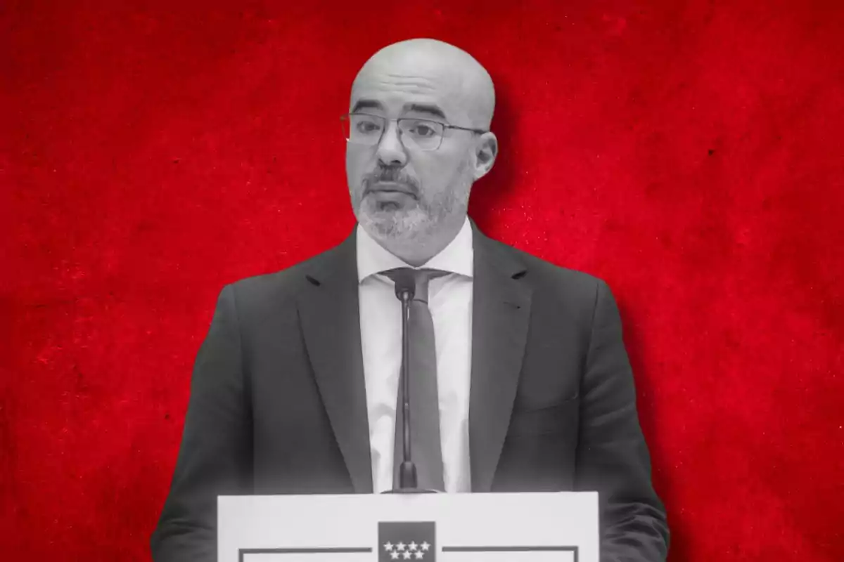 Un hombre con gafas y barba, vestido con traje y corbata, habla frente a un micrófono con un fondo rojo.
