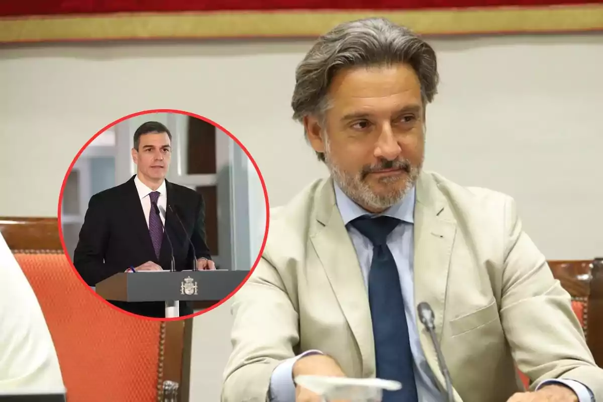 Un hombre con traje claro y corbata azul está sentado en una mesa mientras otro hombre en un recuadro da un discurso.