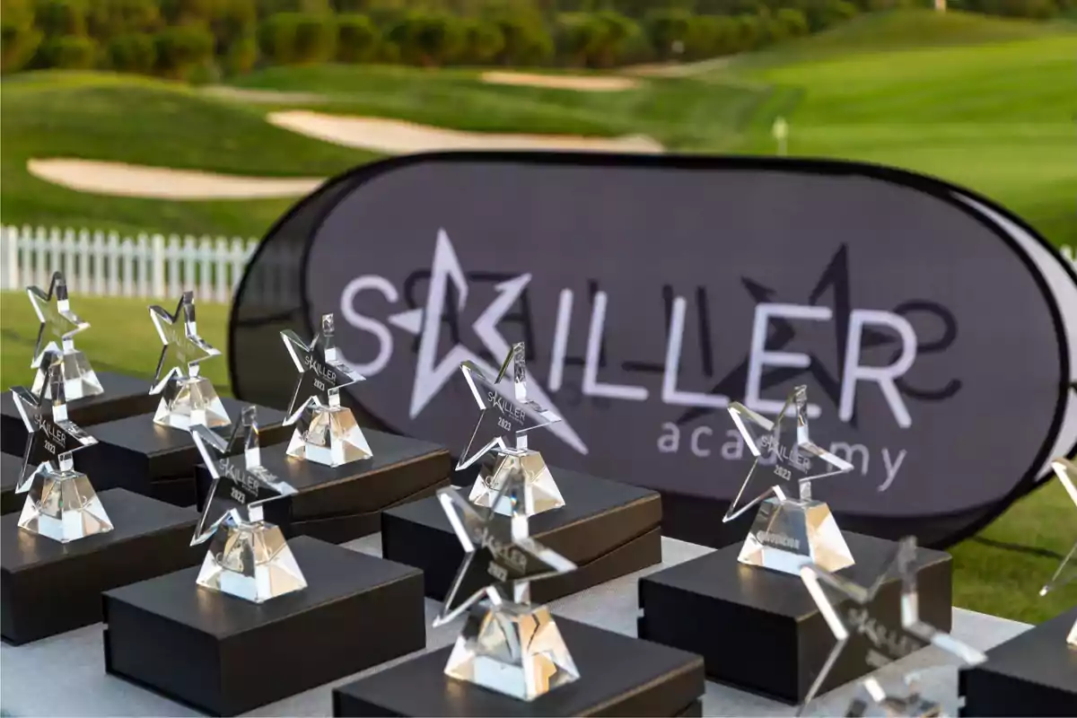 Trofeos en forma de estrella sobre cajas negras con un cartel de fondo que dice "Skiller Academy" en un campo de golf.