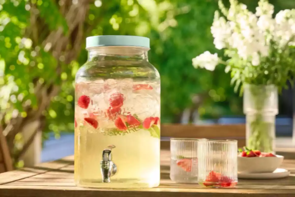 Jarra grande de vidrio con agua fresca, fresas y hojas de menta sobre una mesa de madera al aire libre junto a dos vasos y un tazón pequeño con fresas, con fondo de plantas verdes y flores blancas. Jarra grande de vidrio con agua fresca, fresas y hojas de menta sobre una mesa de madera al aire libre junto a dos vasos y un tazón pequeño con fresas, con fondo de plantas verdes y flores blancas.