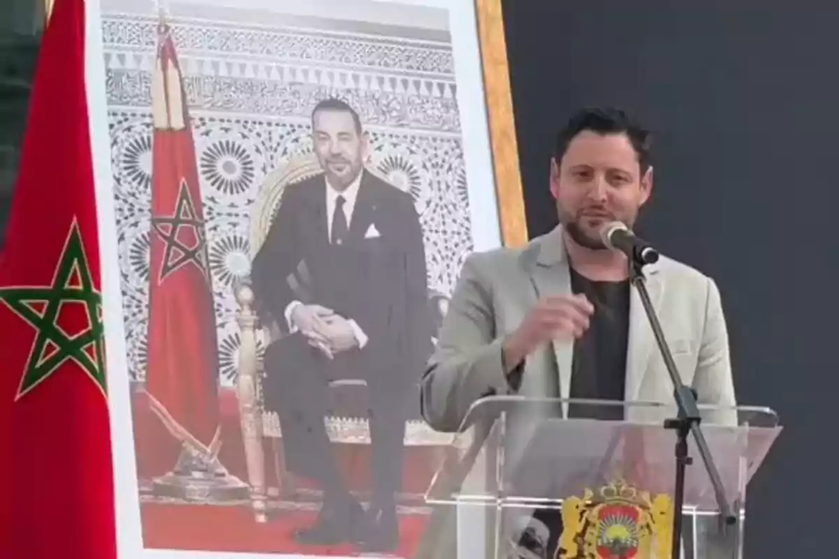 Un hombre habla en un podio con un micrófono frente a una bandera de Marruecos y un retrato de una persona sentada en un trono. Un hombre habla en un podio con un micrófono frente a una bandera de Marruecos y un retrato de una persona sentada en un trono.