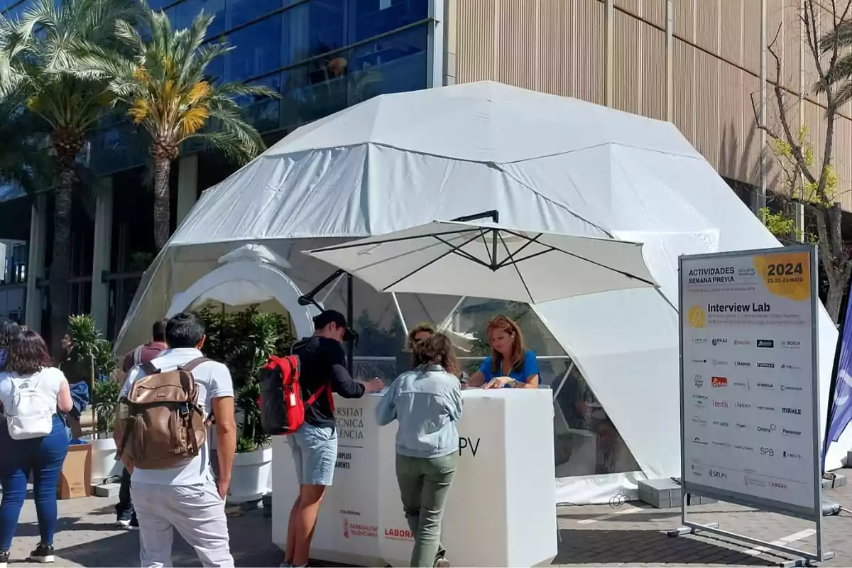 Personas reunidas frente a un stand informativo al aire libre con una estructura geodésica blanca de fondo y un cartel de actividades.