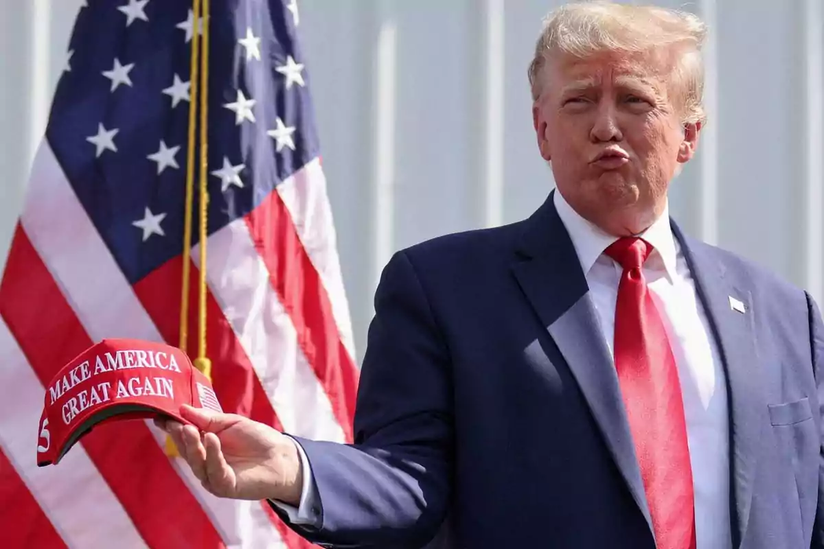 Un hombre con traje azul y corbata roja sostiene una gorra roja frente a una bandera de Estados Unidos.