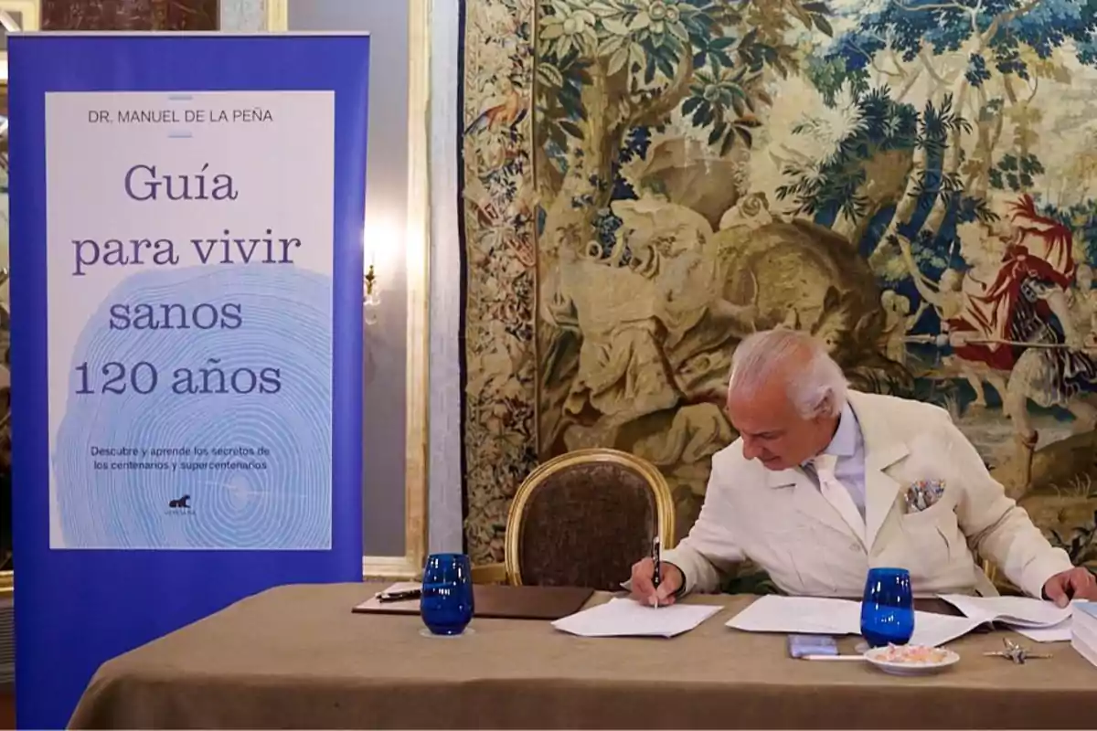 Un hombre de cabello canoso y traje claro está sentado en una mesa firmando documentos junto a un cartel que promociona un libro titulado "Guía para vivir sanos 120 años" de Dr. Manuel de la Peña.