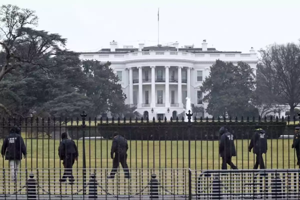 Un grupo de personas camina frente a una valla con la Casa Blanca al fondo en un día nublado.