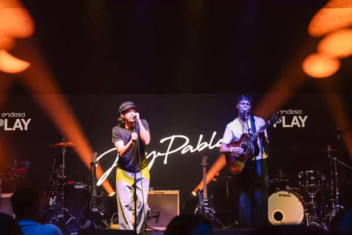 Dos músicos actuando en el escenario con guitarras y batería, luces naranjas y fondo con el nombre de la banda Dos músicos actuando en el escenario con guitarras y batería, luces naranjas y fondo con el nombre de la banda