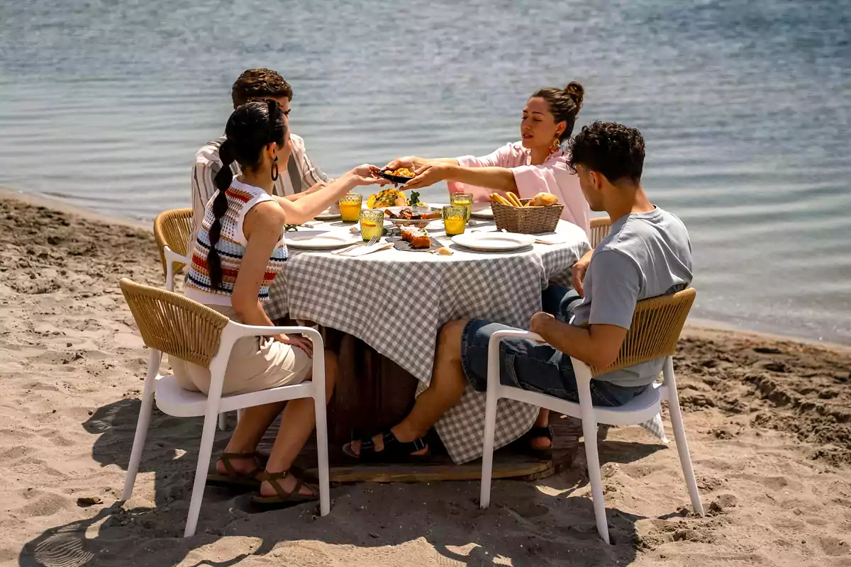 Cuatro personas disfrutan de una comida al aire libre sentados alrededor de una mesa en la playa junto al mar
