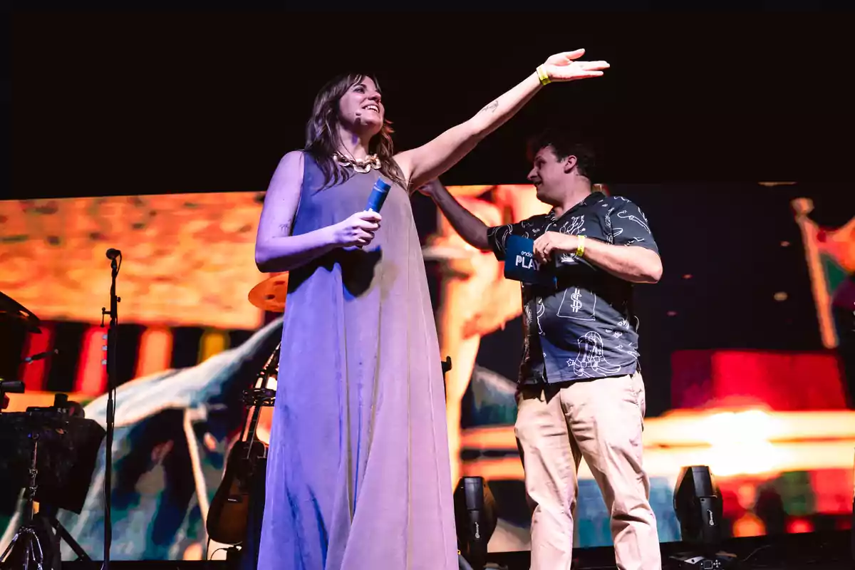 Una mujer sonriente con un micrófono azul levanta el brazo mientras un hombre a su lado sostiene una tarjeta y la mira en un escenario iluminado Una mujer sonriente con un micrófono azul levanta el brazo mientras un hombre a su lado sostiene una tarjeta y la mira en un escenario iluminado