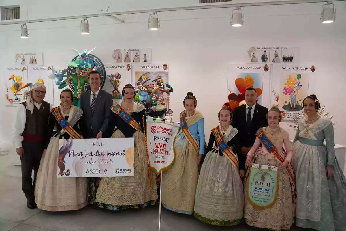 A group of people dressed in traditional costumes pose at an award event, holding a check and a banner, surrounded by colorful figures and fallas posters.