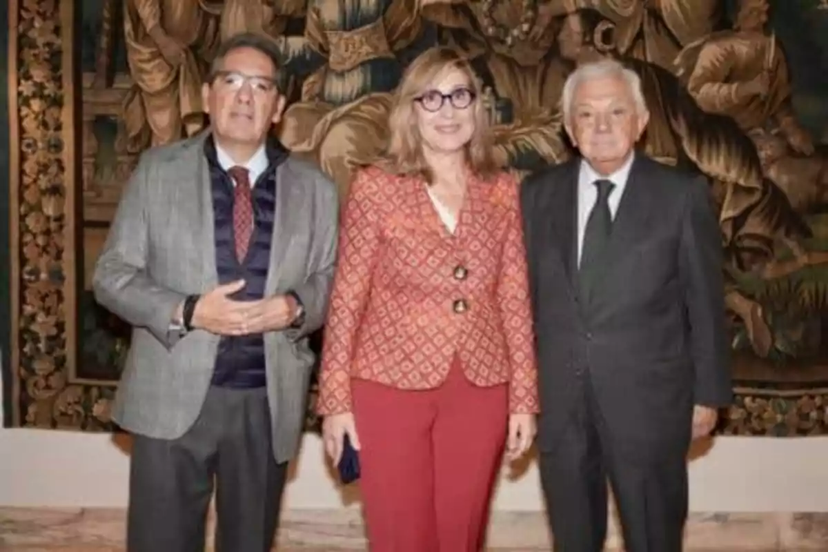 Tres personas posan frente a un tapiz decorativo.