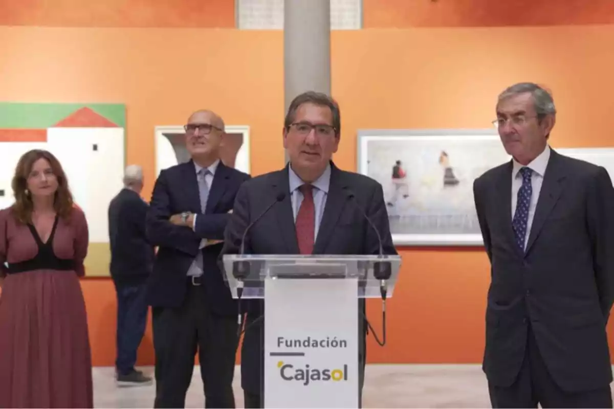 Un grupo de personas en un evento de la Fundación Cajasol, con un hombre hablando en un podio frente a una pared naranja con obras de arte.