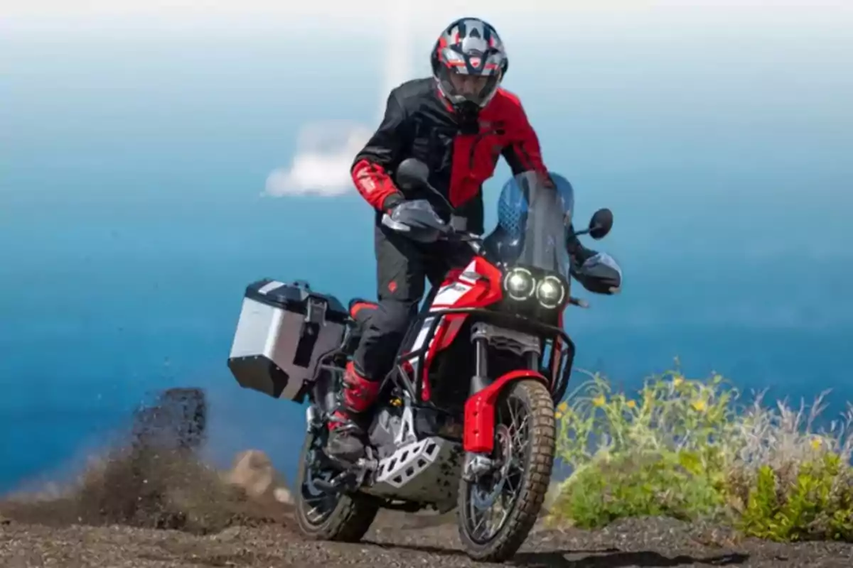 Persona conduciendo una motocicleta todoterreno roja en un camino de tierra con el mar de fondo.