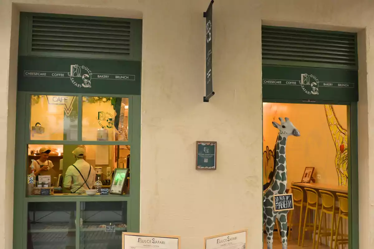 Fachada de una cafetería con ventanas y puertas verdes, en el interior se observa una jirafa decorativa y personas atendiendo el mostrador
