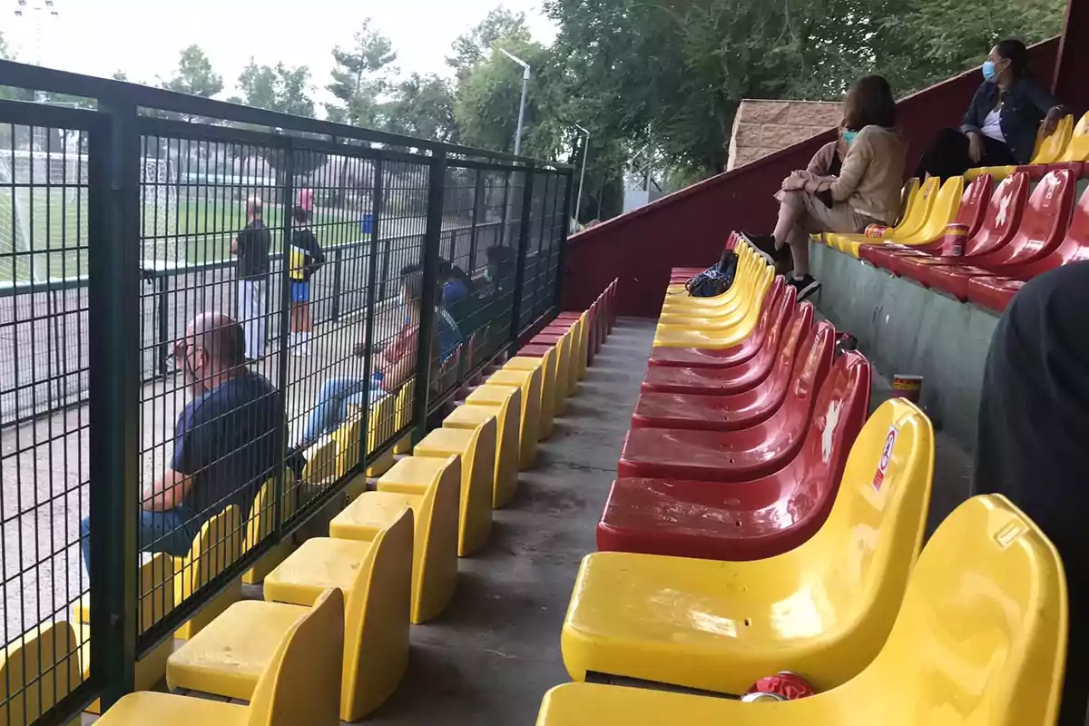 Gradas de un estadio con asientos amarillos y rojos, algunas personas sentadas y otras de pie observando un campo de fútbol a través de una reja.