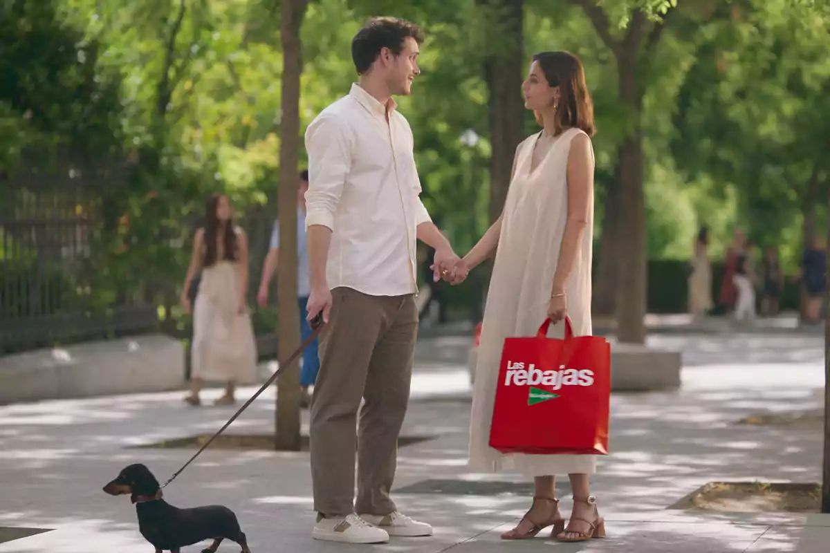 Una pareja se encuentra en un parque, sosteniéndose de la mano mientras pasean a su perro, y la mujer lleva una bolsa roja que dice "Las rebajas".