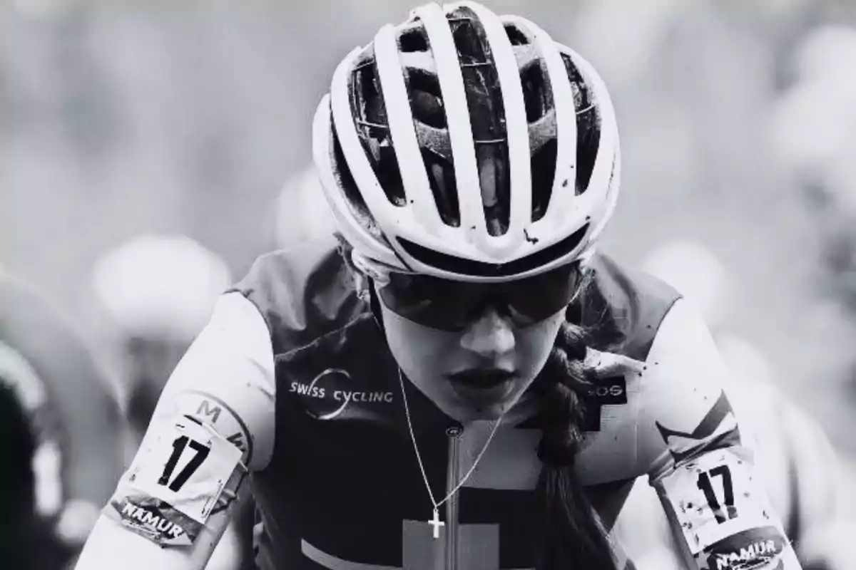 Ciclista con casco y gafas de sol en una carrera, lleva el número 17 en el brazo y un collar con una cruz.