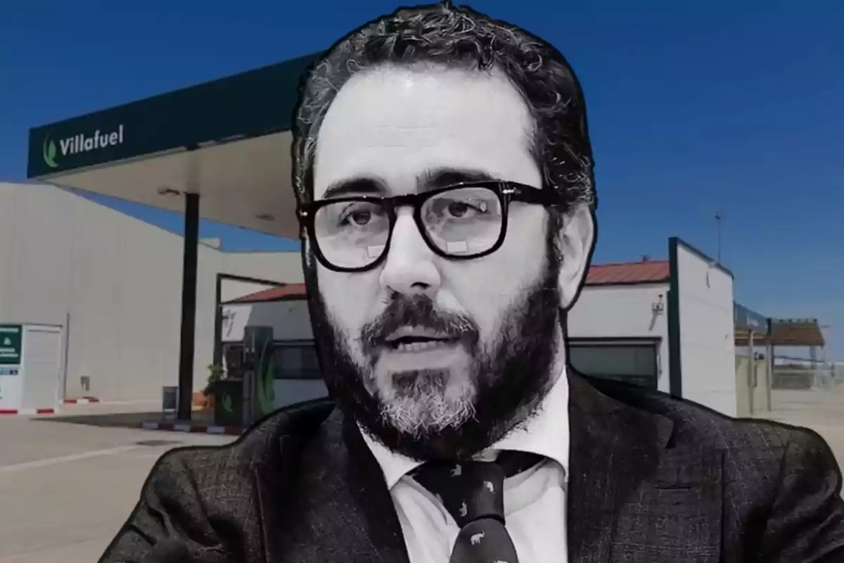 Un hombre con barba y gafas frente a una estación de servicio Villafuel.