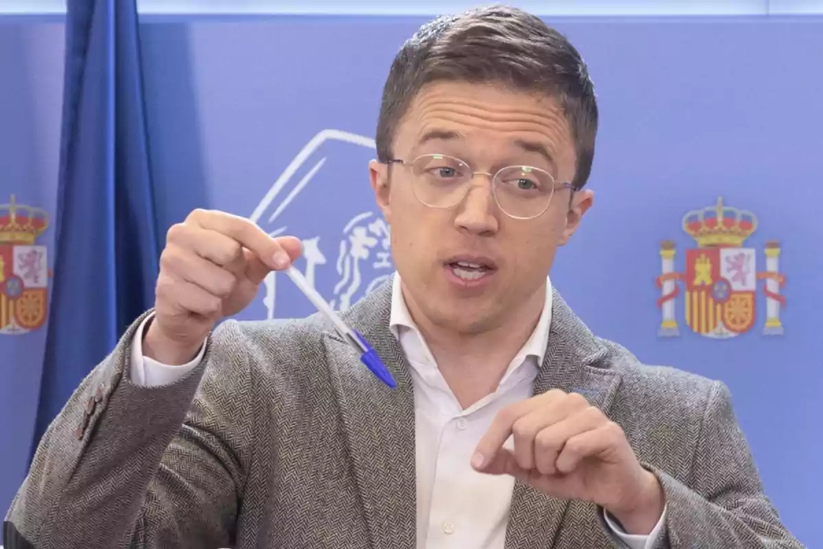 Un hombre con gafas y traje sostiene un bolígrafo mientras habla frente a un fondo con banderas de España.