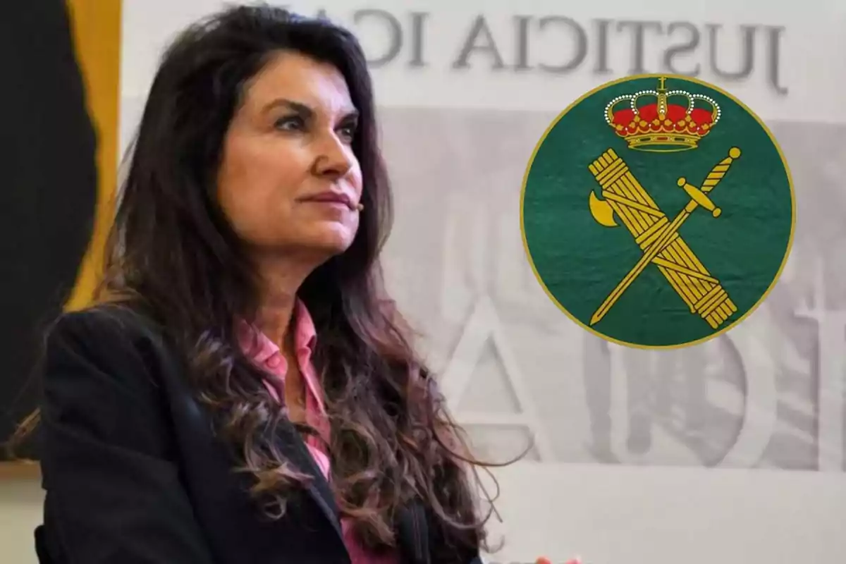 Mujer de cabello oscuro con chaqueta negra y camisa rosa sentada junto a un emblema circular verde con una corona y símbolos dorados.